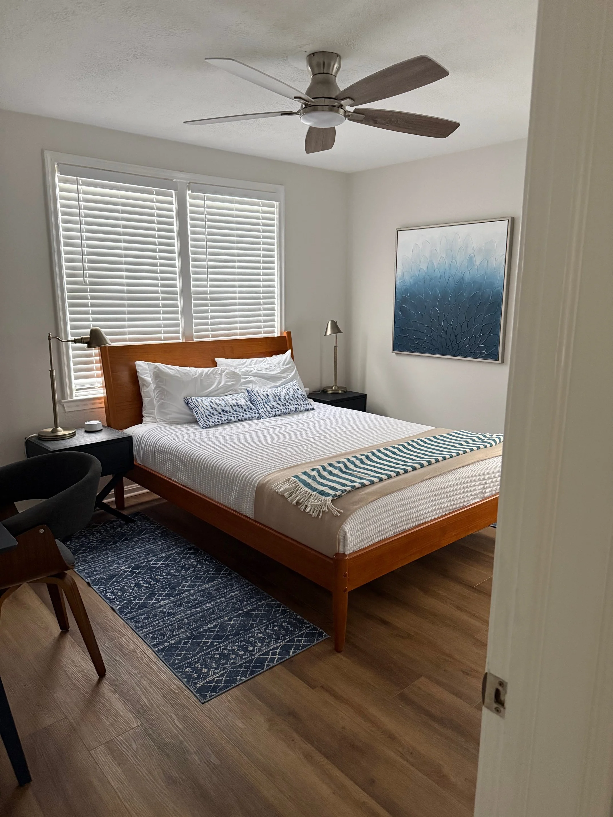 A cozy bedroom with a wooden bed frame, white bedding, and blue decorative pillows. There are two black nightstands with lamps, a window with white blinds, a blue abstract painting on the wall, a ceiling fan, and a patterned blue rug on wood flooring