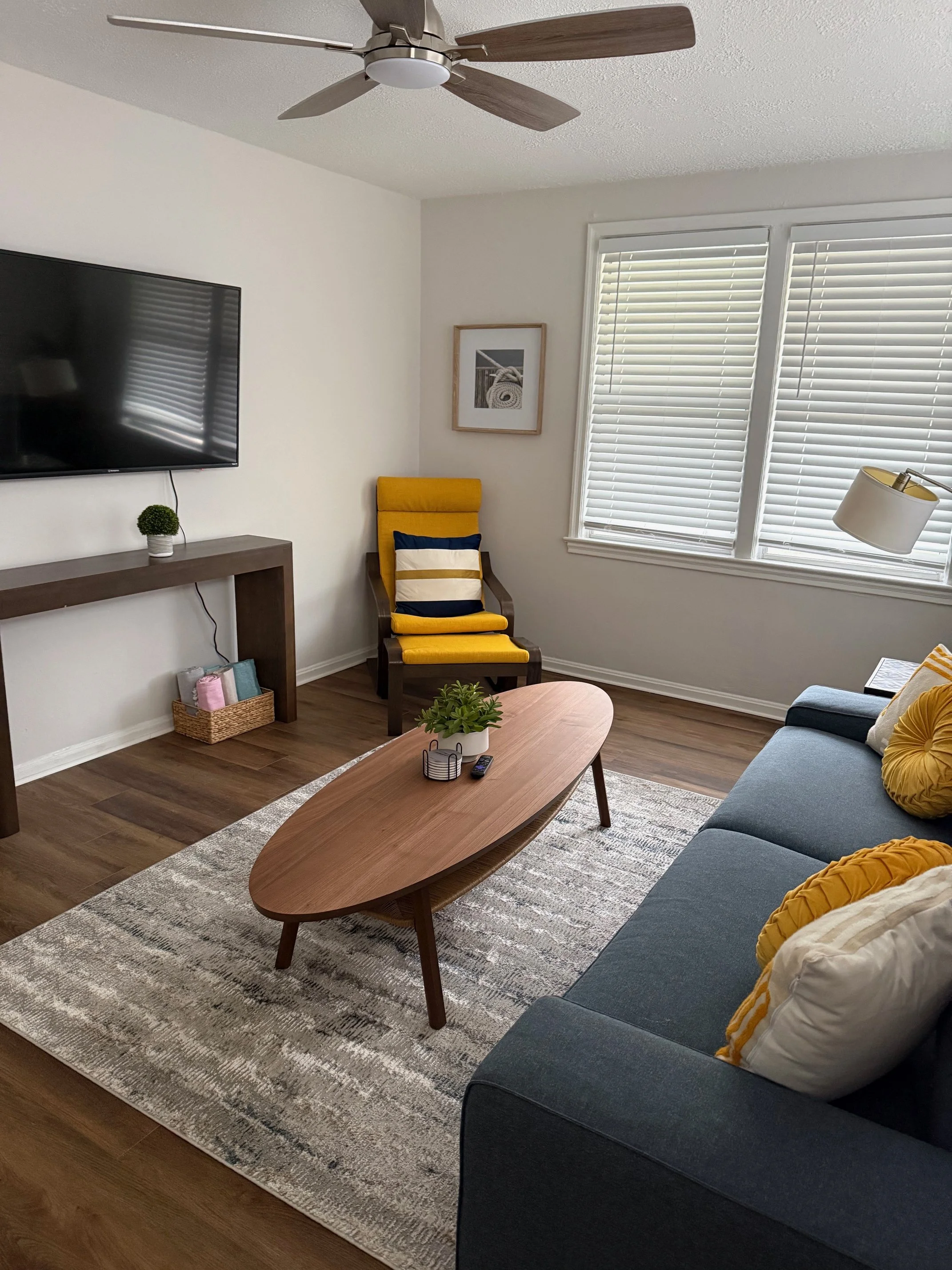 Living room with a blue sofa with yellow pillows, a wooden coffee table with a plant, a gray rug, a yellow and gray armchair, a wall-mounted TV, window blinds, a floor lamp, and wall art.