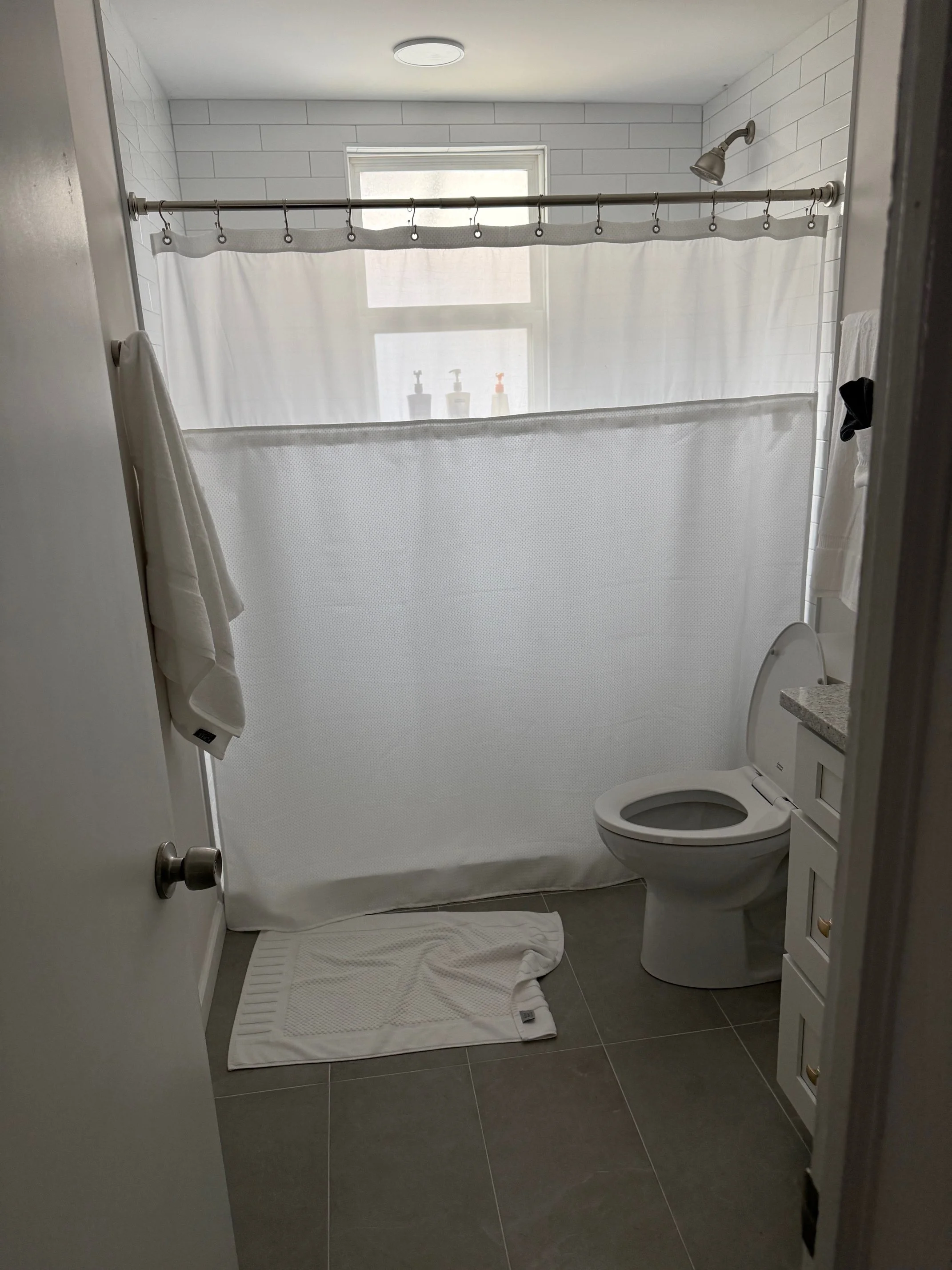 A small bathroom with a white shower curtain, a toilet, a white towel hanging on the wall, a bath mat on the floor, and a bathroom vanity with a granite countertop. There are bottles on the window sill and a window with frosted glass.