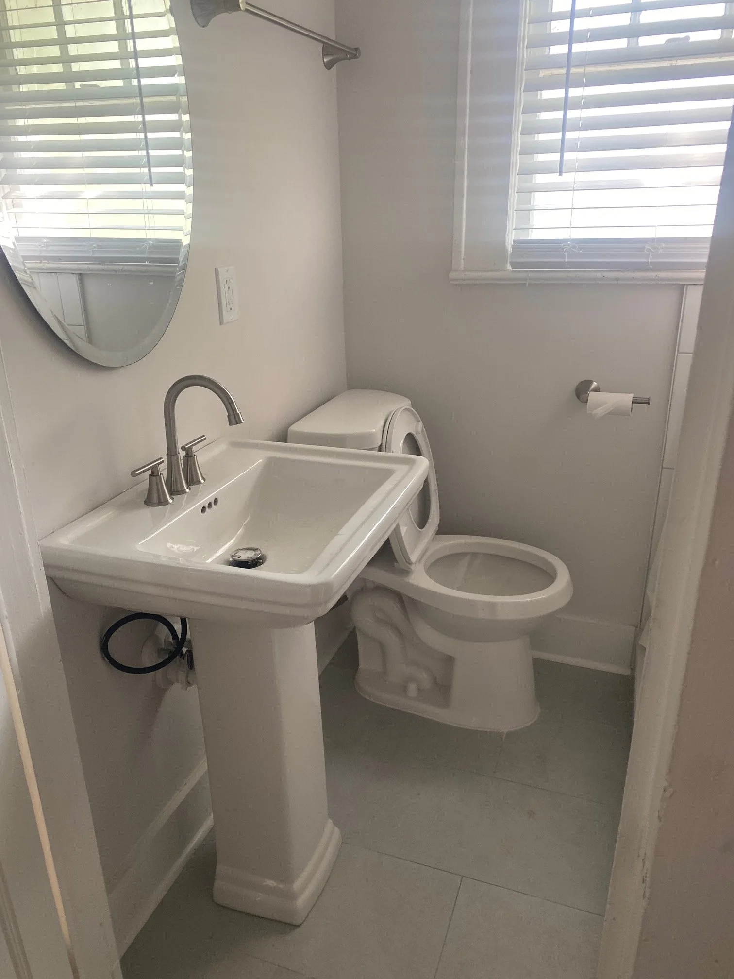 A small bathroom with a white pedestal sink, a mirror, a toilet, and two windows with white blinds.