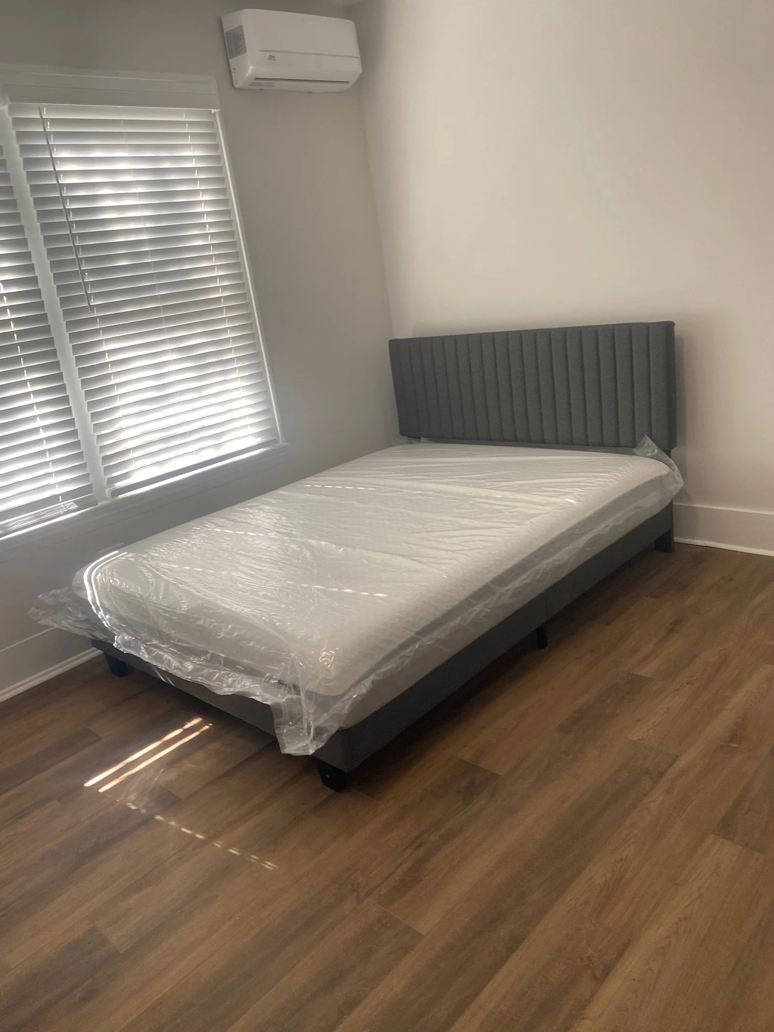 An empty bedroom featuring a bed with a gray, vertical striped headboard and a plastic-covered mattress. The room has a window with closed white blinds and a wall-mounted air conditioner.
