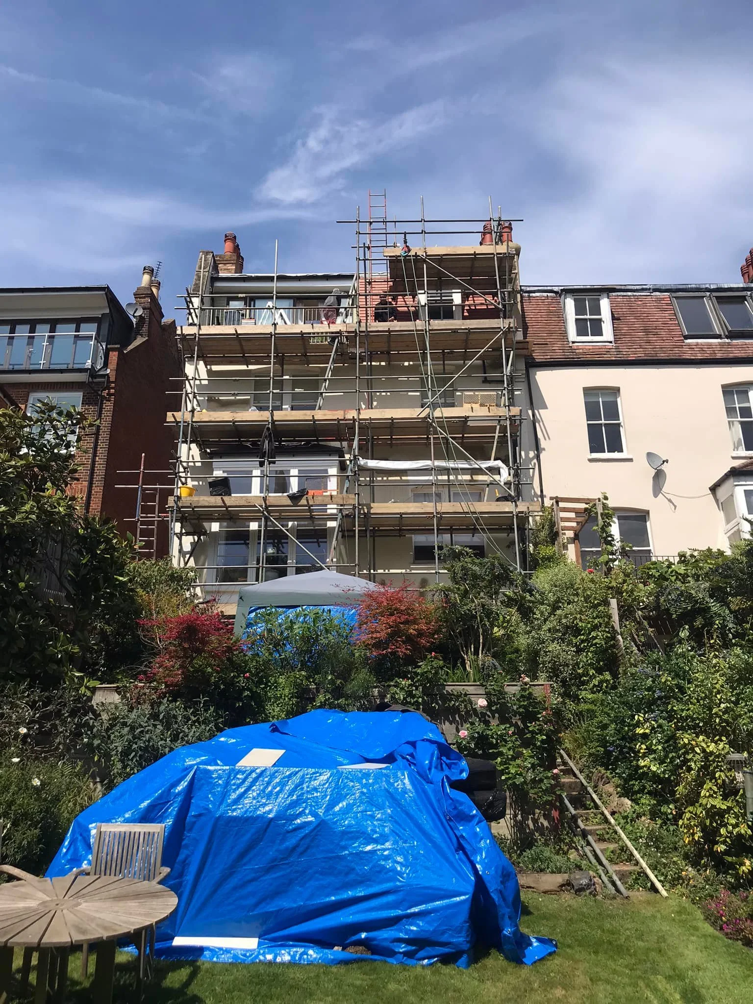 Building with scaffolding, gardening tools, and equipment in a residential area with a blue tarp covering some items in the foreground.
