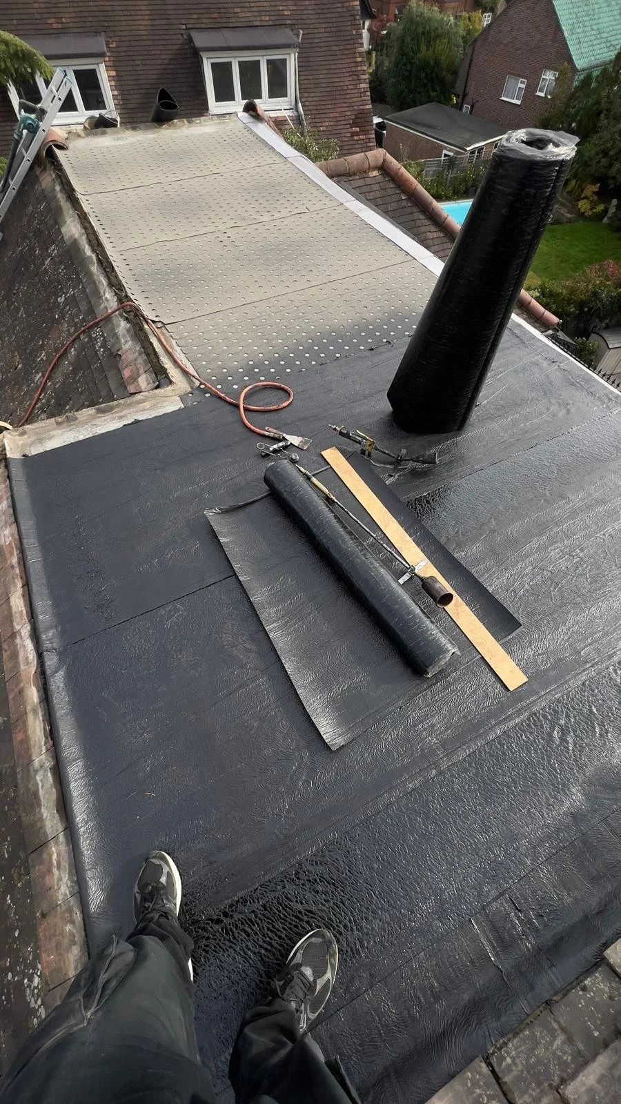 Person working on roof in North London for K G Croft with black waterproofing membrane, black vent pipe, and tools, in a residential neighbourhood.