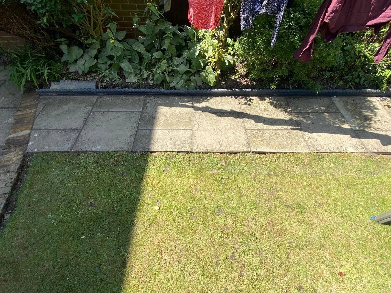 A garden border with green plants and a hanging laundry drying rack with clothes, next to a paved stone pathway, with grass in the foreground.
