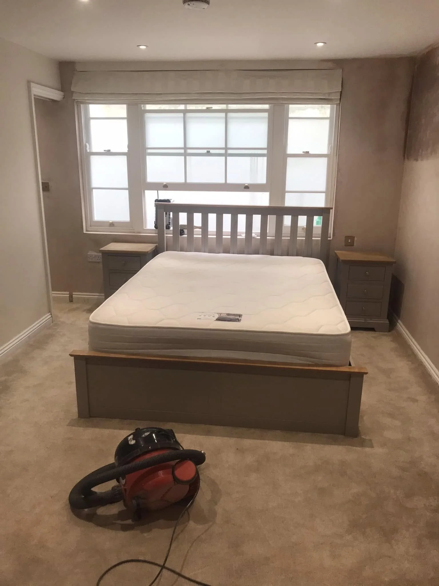 Empty bedroom with a bed and a vacuum cleaner on beige carpet.