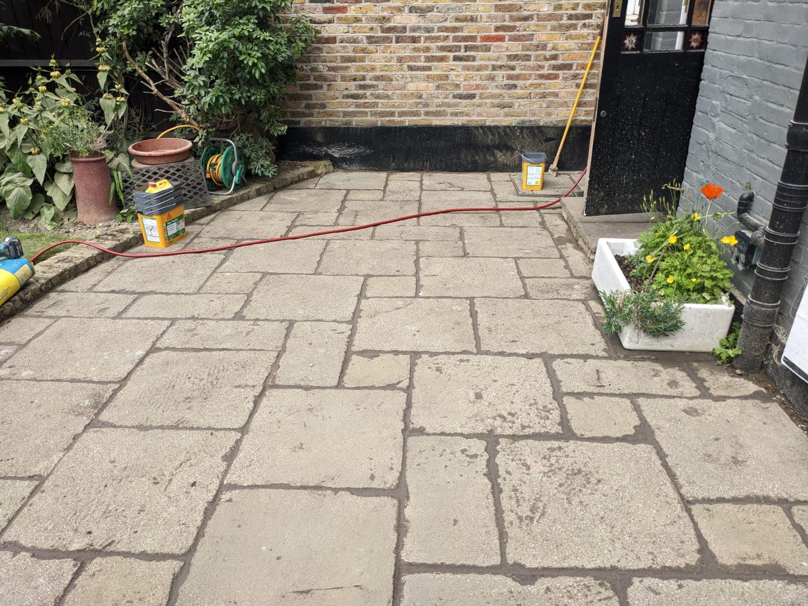 A paved outdoor patio with large square stone tiles, surrounded by plants and gardening tools, with a brick house wall in the background.