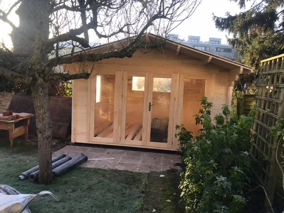 New wooden shed in a backyard, surrounded by trees, plants, and a garden fence.