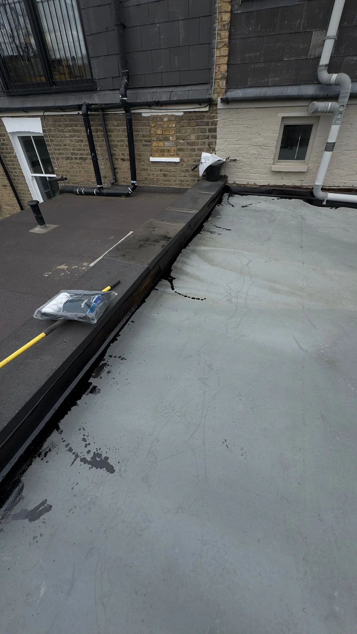 A rooftop in north london with partially installed waterproofing membrane, black drainage system, and various tools including a roller and a bag of waterproofing material. The image shows the edge of the roof with a wall and pipes in the background.
