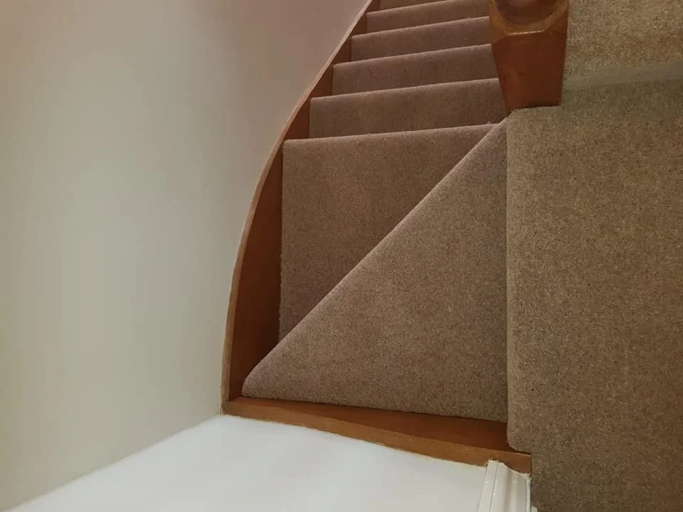 Carpeted staircase with beige carpeting, curved wooden railing on the right side, and a beige wall on the left side.