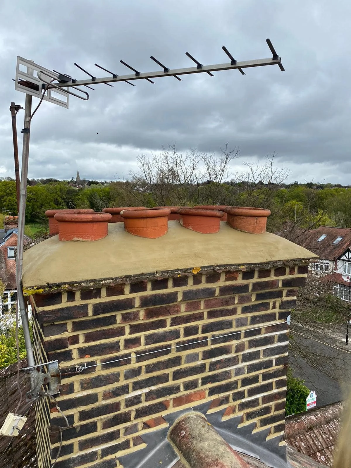 View of a chimney with a brick exterior and a light-colored chimney cap, topped with multiple terracotta chimney pots. An outdoor TV antenna is mounted on the chimney with a cable connected to it. The background shows a cloudy sky and a distant lands