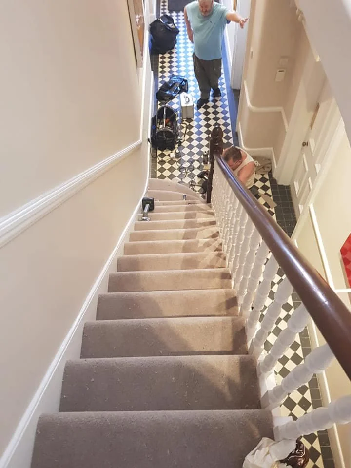 A top-down view of a staircase looking down to a hallway with two men standing at the bottom. One man is pointing at something. The hallway has black and white checkered flooring, with various items scattered around, including bags, boxes, and a vacu