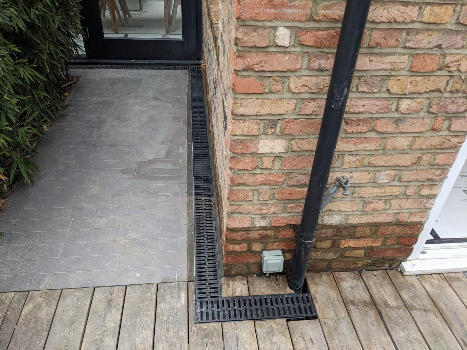 Exterior corner of a building with brick wall, black drain pipe, and a water spigot near ground. Floor consists of wooden deck and gray tiles.