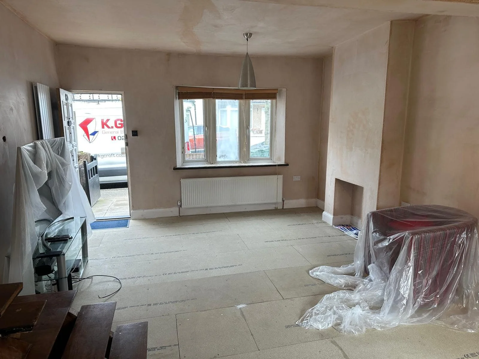 Empty living room with beige walls, a window with blinds, a radiator, and furniture covered in plastic. The floor is unfinished, and there's an open door leading outside.