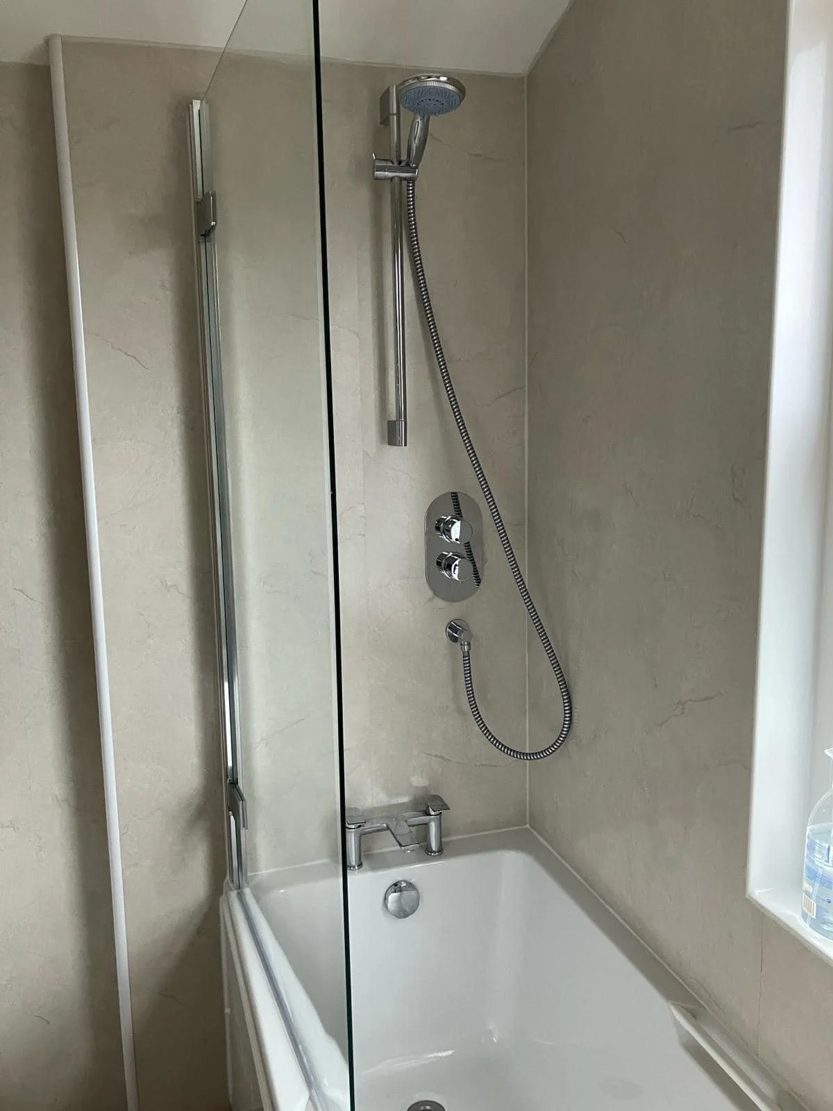 Bathroom shower with beige tiled walls, a glass shower door, a chrome handheld showerhead, and a built-in shelf with a bottle of water.