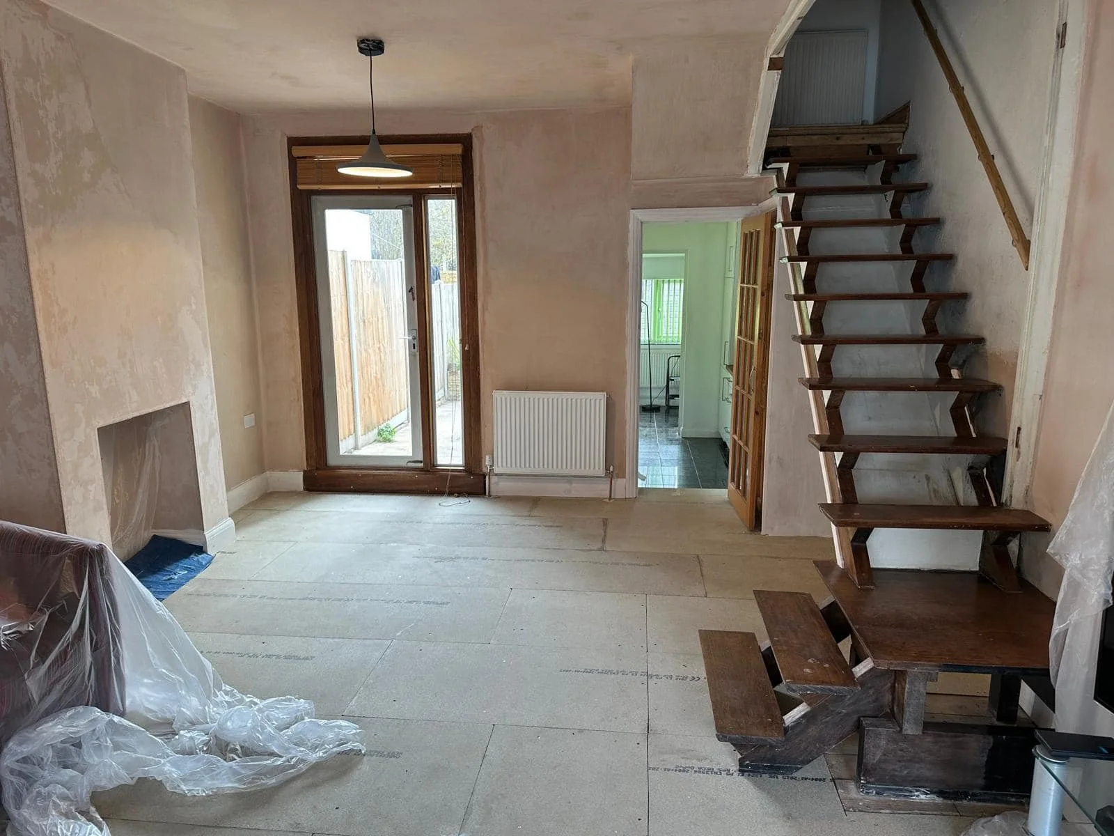 Living room in north london undergoing renovation with unfinished walls, plastic sheet on the couch, a sliding glass door leading outside, a staircase with unfinished steps, and a doorway leading to another room by K G Croft.