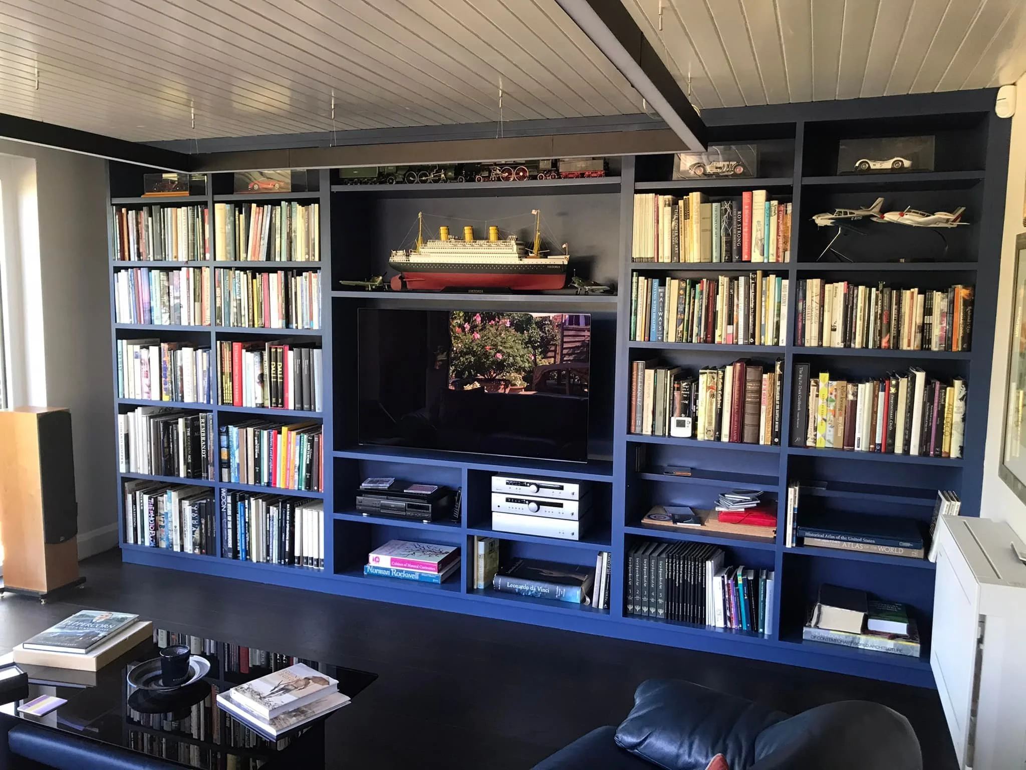 Blue bookshelf made by K G Croft filled with books and model ships, television in the center, and a table with magazines in front. In north london.