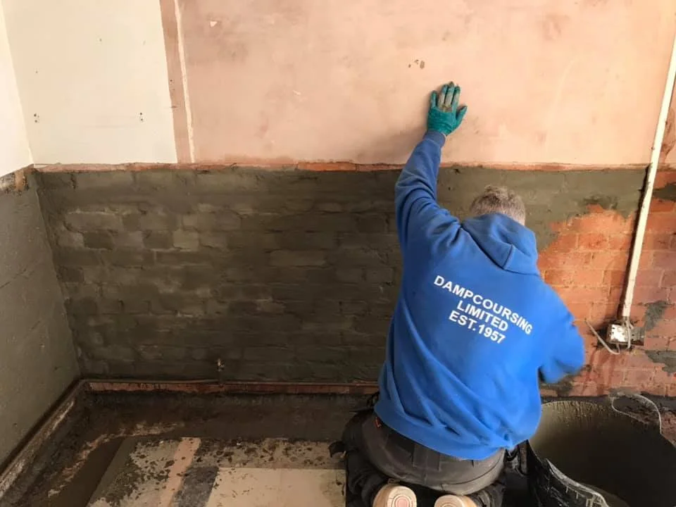 A construction worker kneels on the floor and paints a wall with pink paint. The worker is wearing a blue hoodie with white text on the back that says "DAMP COURSING LIMITED EST. 1957".