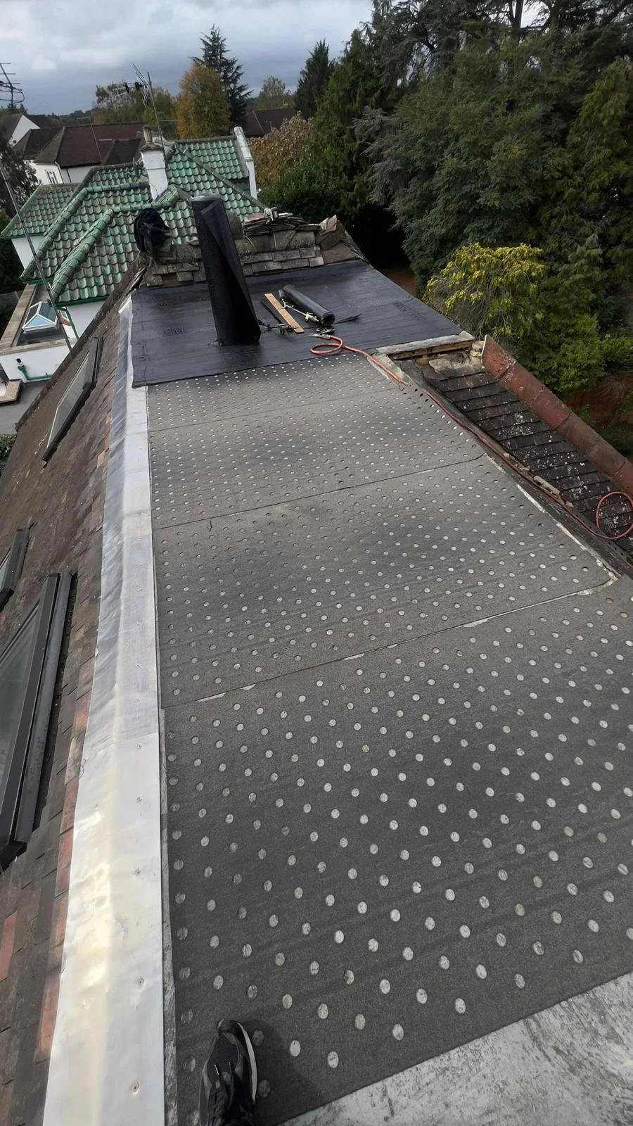 Roof under construction by K G Croft in North London with insulation and roofing materials, tools, and vent pipes, in a residential area with trees and houses in the background.