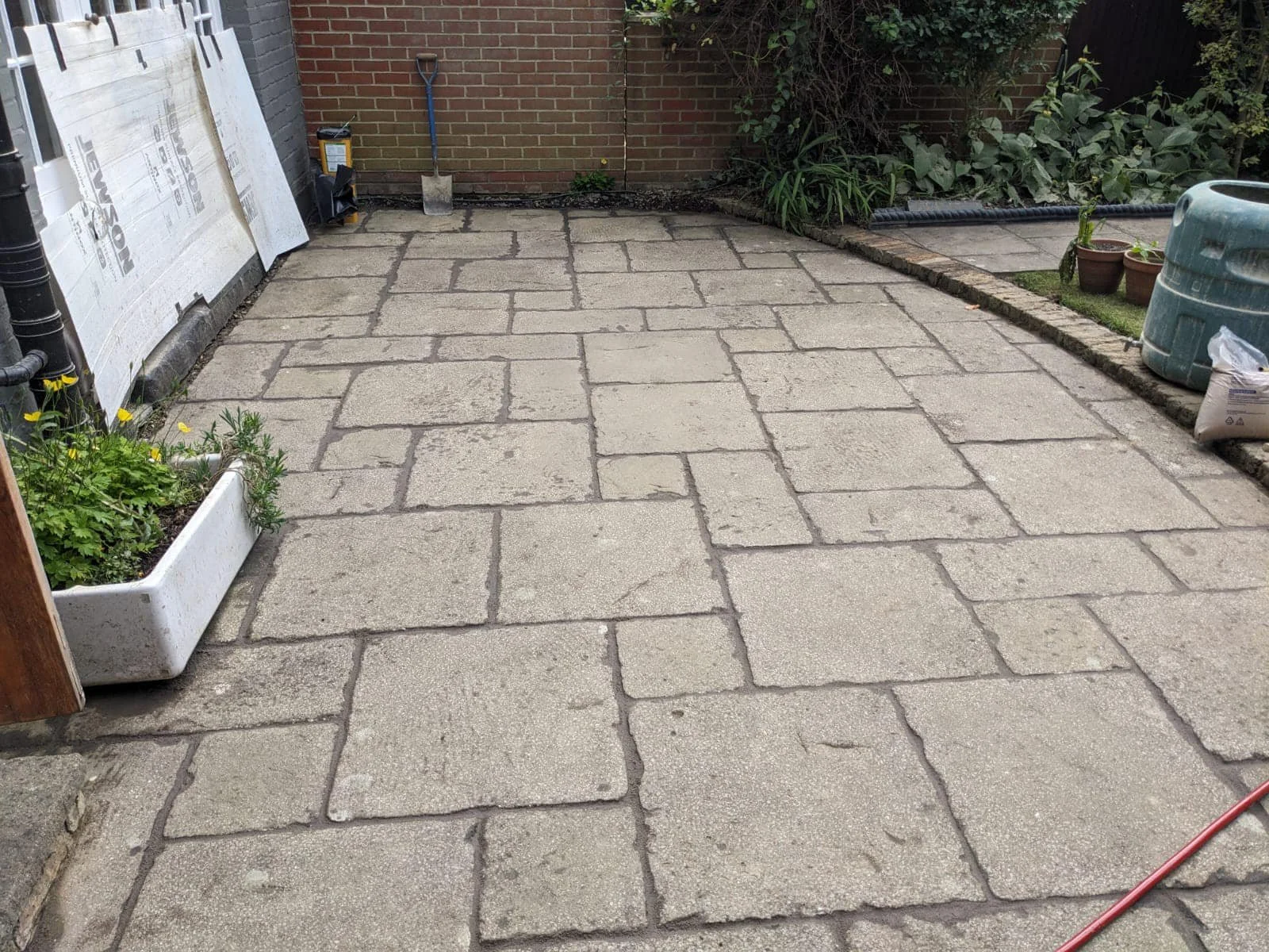 A newly paved stone patio with large rectangular concrete pavers in a backyard, with garden plants in the background and construction tools nearby.