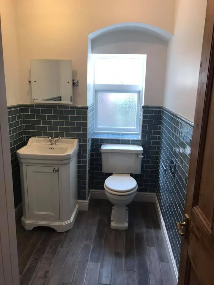 Small bathroom with a toilet and a small white sink, a mirrored cabinet above, a frosted window, and dark green tiled walls halfway up, with a wood floor.