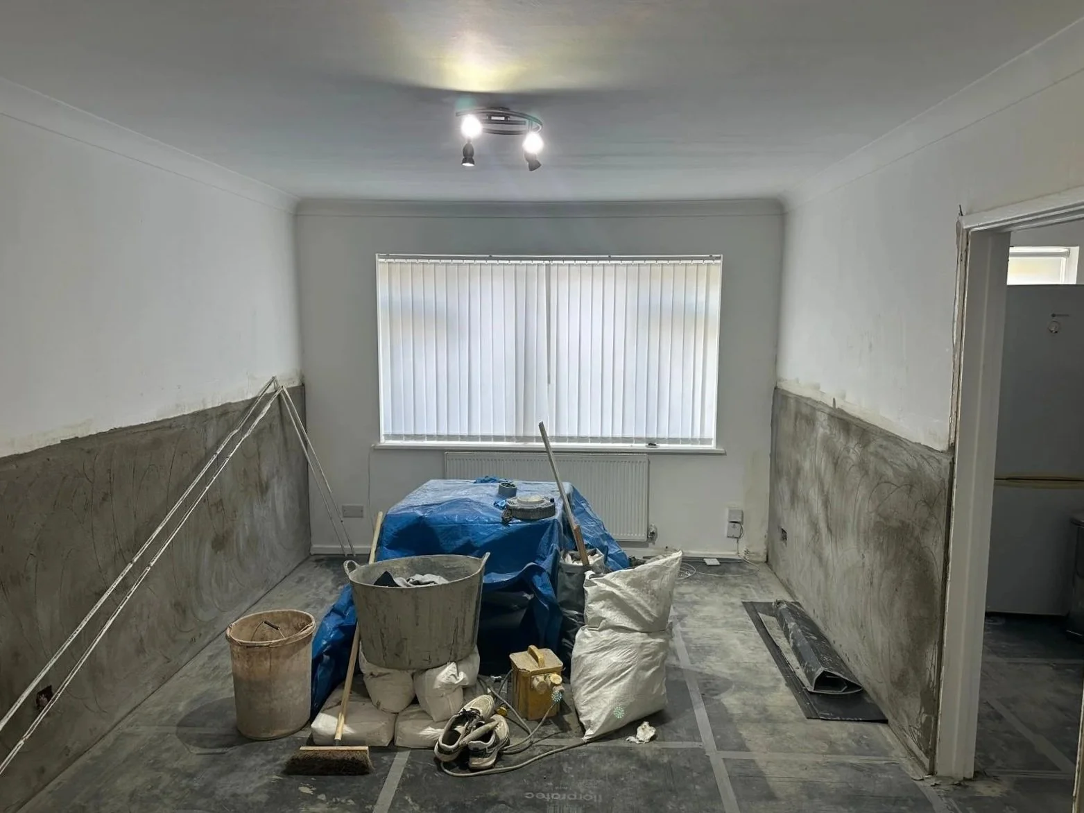 A room under renovation in north london by K G Croft with drywall patches on the lower half of the walls, construction tools, a bucket, bags of materials, a window with closed vertical blinds, and construction tape on the floor.