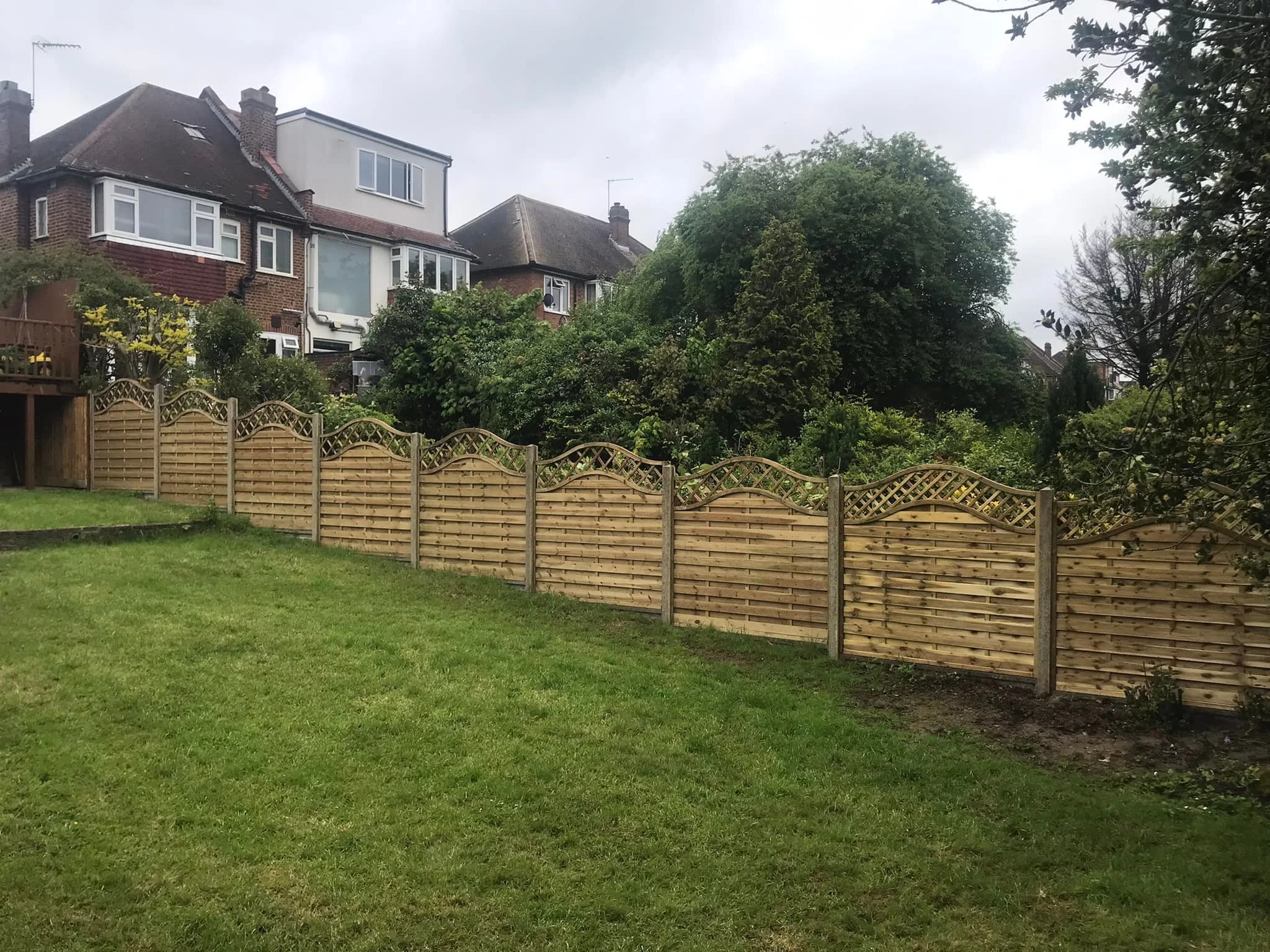 A backyard with green grass and a decorative wooden fence separating it from neighboring houses, with trees and a cloudy sky in the background.