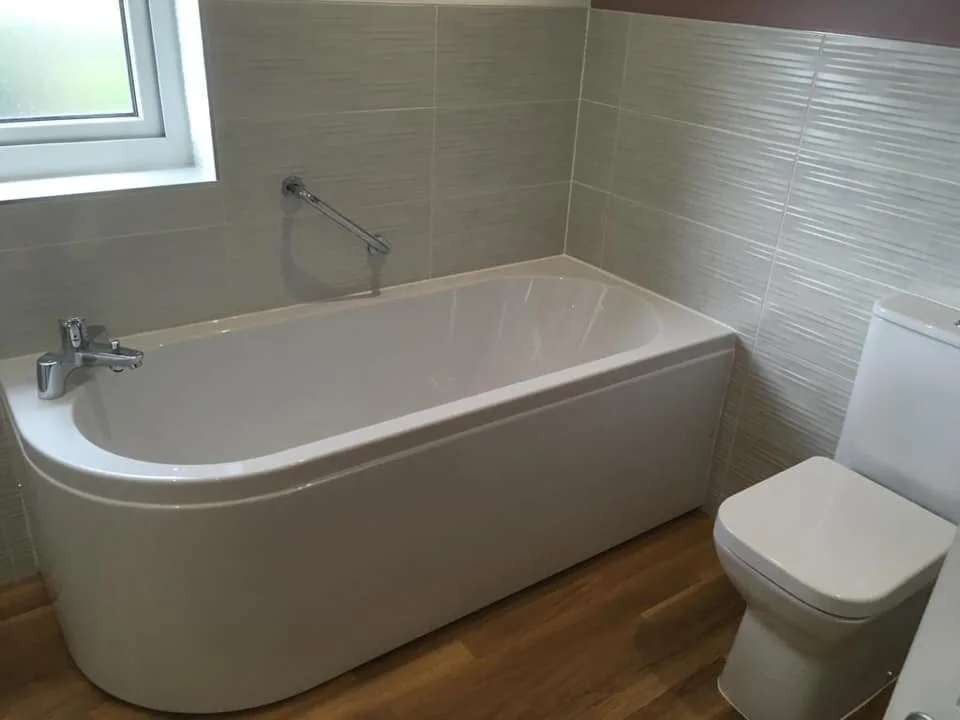 Bathroom with a bathtub, a window, and a toilet, featuring beige tiled walls and wooden floor.