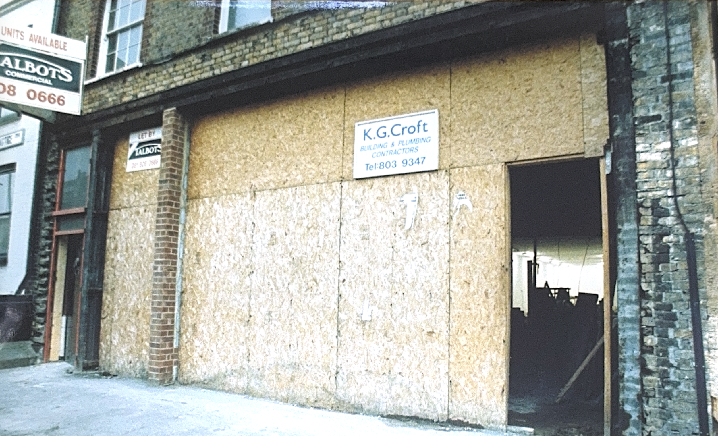 The storefront is boarded up with plywood, with a sign for K.G. Croft building & plumbing contractors, and an unfinished interior visible through an open doorway. Other signs and brick building details are visible.