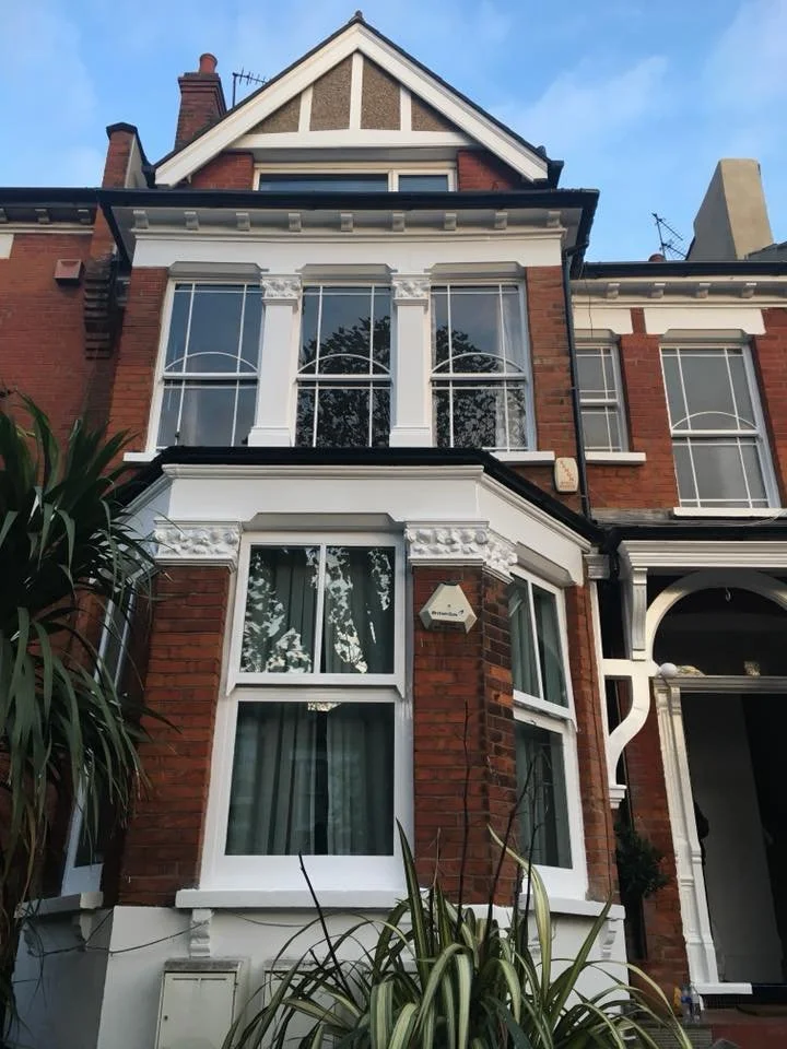 A multi-story brick house in north london with tall, arched windows, decorative white trim, and a steep gabled roof, with plants in the foreground and advertisements of a clear sky.
