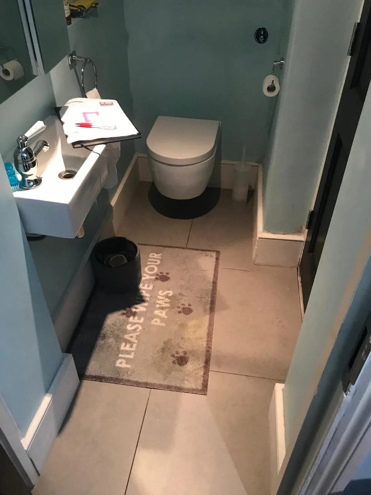 Small bathroom with a toilet, a small sink with a faucet, a black wastebasket, and a rug that says 'Please remove your paws' with paw prints. There are some papers and pens on the sink and a roll of toilet paper on a holder.