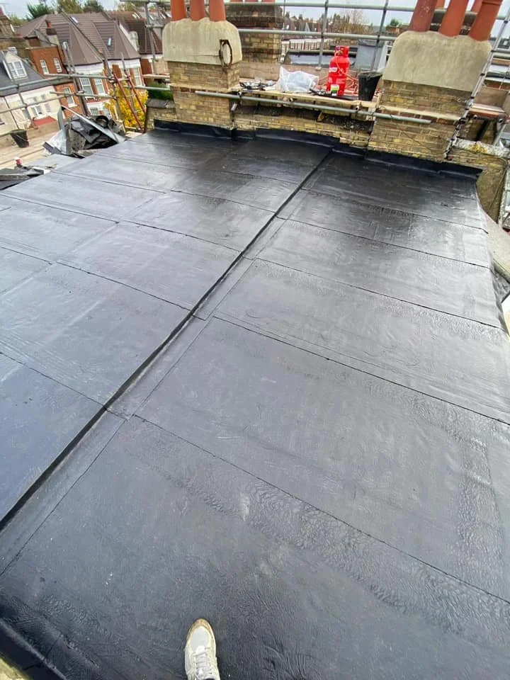 View of a flat rooftop covered with new black waterproofing membrane, chimney stacks in the background, construction tools and materials on the chimney ledge, and a person's foot visible at the bottom of the photo. K G Croft.