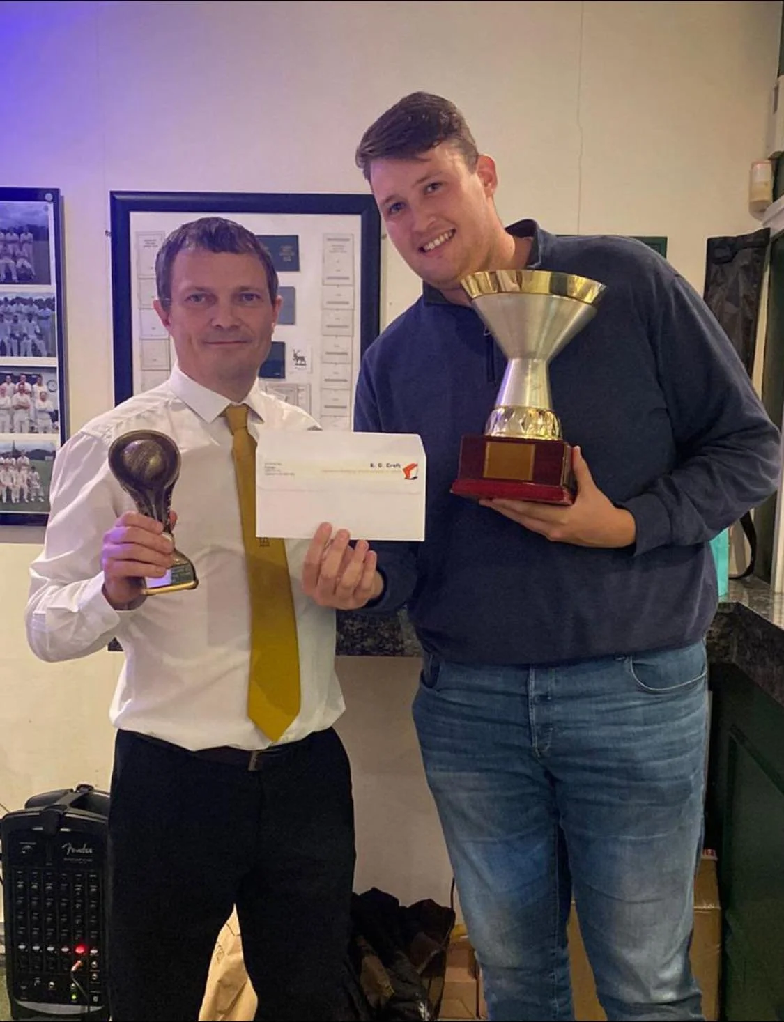 Two men of Southgate Adelaide cricket club standing side by side, holding trophies and a check, smiling. One is dressed in a white shirt and yellow tie, and the other in a dark shirt and jeans. Sponsored by K G Croft.