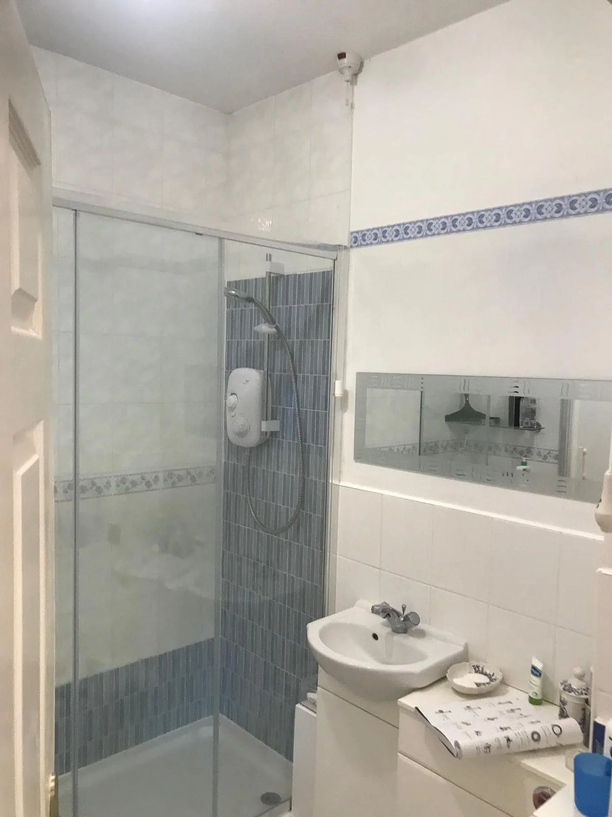 Small bathroom with a shower stall, white sink, mirror, and various toiletries on a counter.
