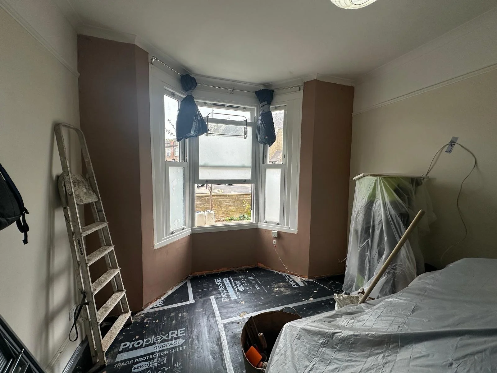 A room under renovation with a bed covered in plastic, a ladder leaning against the wall, and a window with black bags hanging from a curtain rod. The floor is covered with a black protective sheet, and there is a plastic-wrapped piece of furniture i
