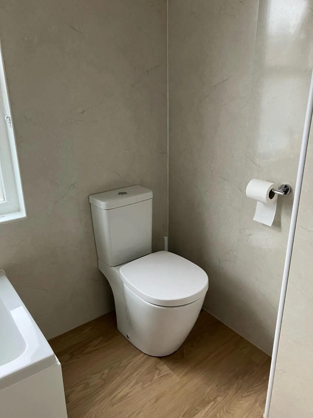 A clean and modern bathroom with a white toilet, beige marble walls, a small window, and a wooden floor.