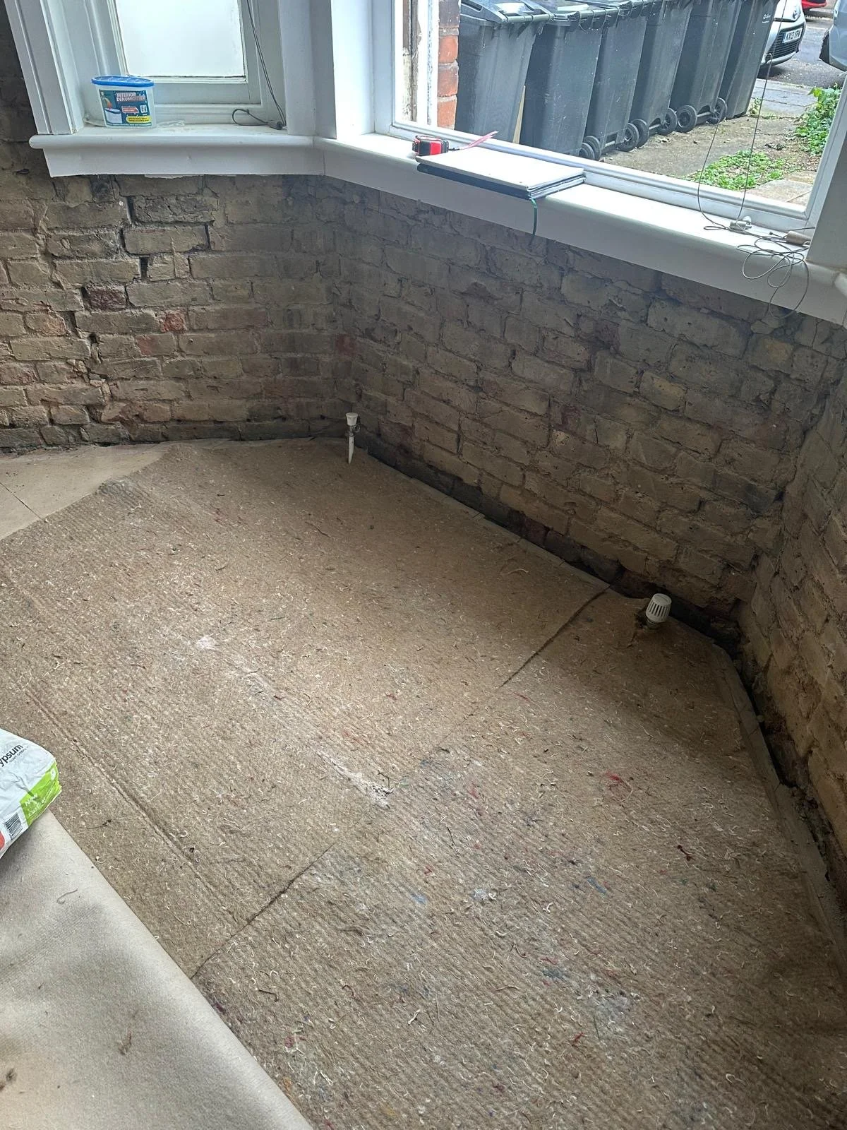 A room under renovation with exposed brick walls, a partially installed carpet, and a window sill with construction tools. Outside, there are trash bins and parked cars.