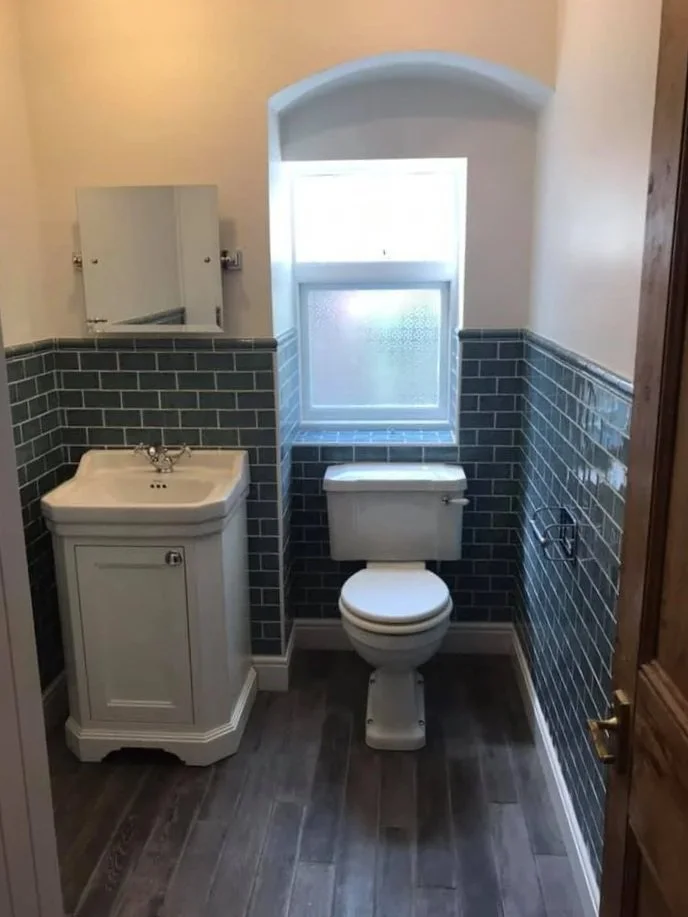 Small bathroom with dark grey tile walls, a small white sink on the left, and a white toilet beneath a window. The floor has wood-like tiles, and a wooden door is partially visible on the right. North London building contractors K G Croft.