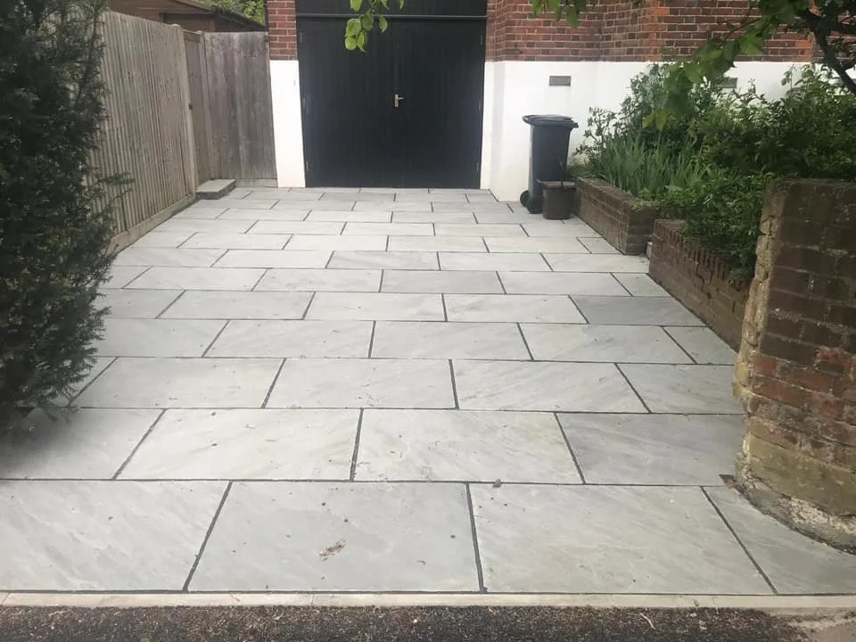 A paved front yard with large rectangular light grey stone tiles, a wooden fence on the left, a black gate in the center, and brick wall on the right with greenery. There is a trash bin and a small container near the gate. K G Croft patio and drive.