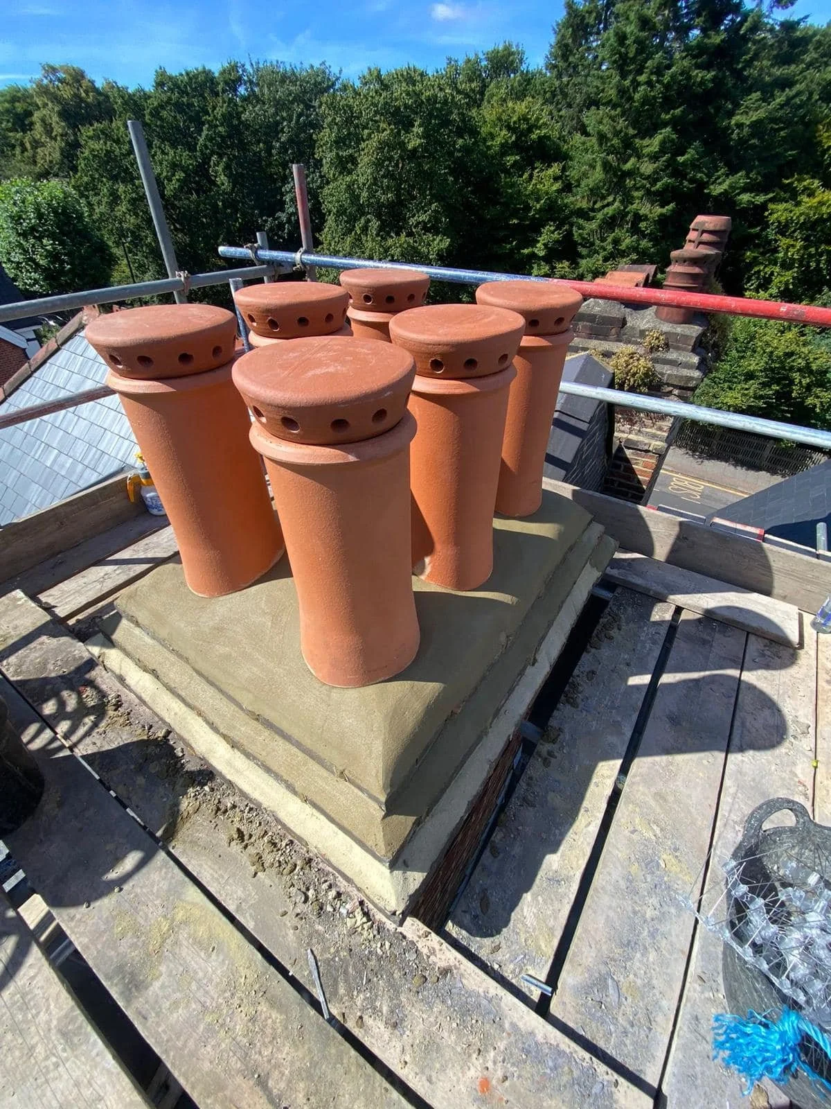 On a rooftop in north london, several new clay chimney pots are stacked together, with scaffolding and safety railing visible, and a background of green trees and a blue sky.