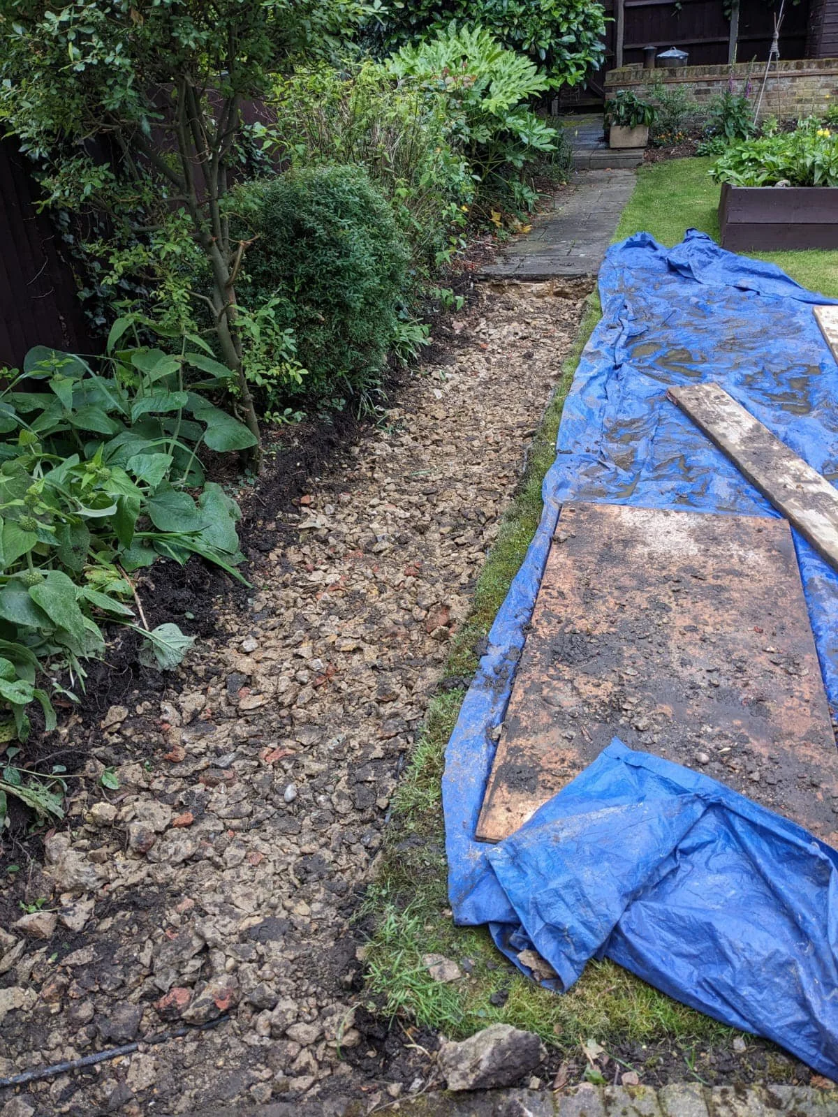 Backyard garden showing a pathway being renovated with gravel, covered by a blue tarp, stones, and wooden planks. K G Croft of north london building contractors.