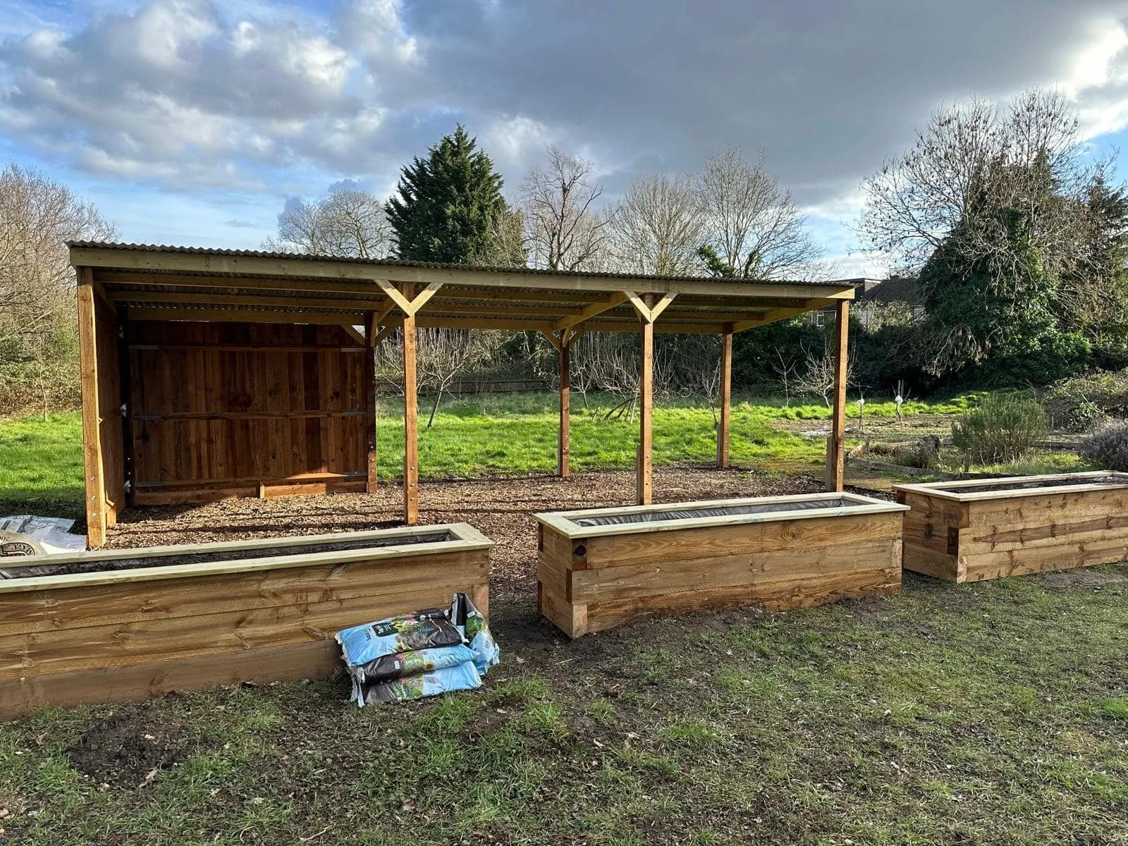 A backyard garden in north london with a wooden raised bed and a wooden shelter or shed under construction, with gardening supplies in the foreground and trees and cloudy sky in the background. Works by K G Croft.