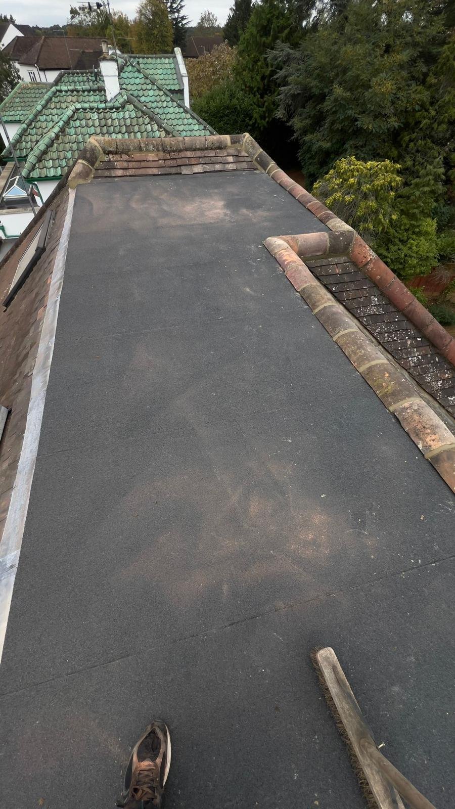 View from the roof in north london showing black flat roofing material, with a broom and a shoe in the foreground, and neighbouring rooftops and trees in the background. K G Croft roof renovation.