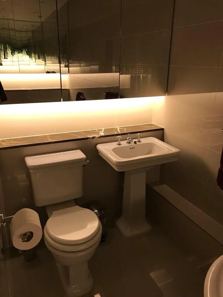 A small bathroom with a toilet and pedestal sink, a large mirror, and dim lighting from behind the mirror, creating a modern and sleek ambiance.