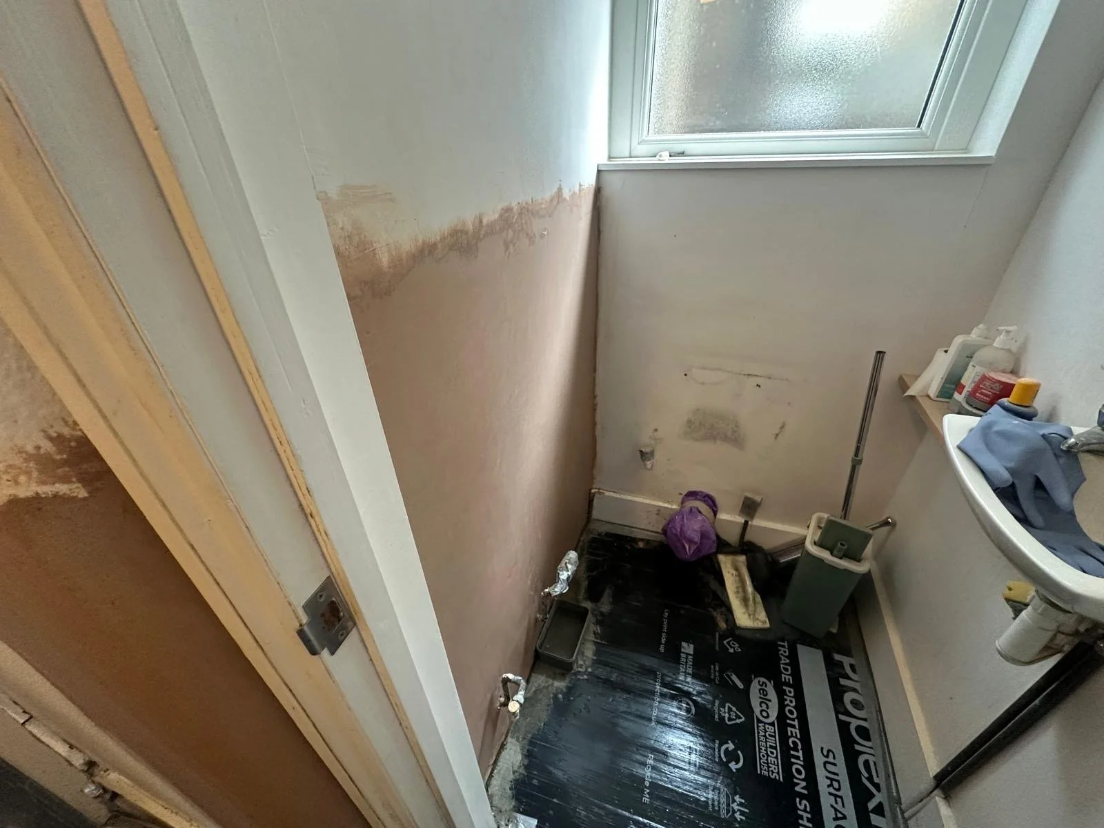 A small bathroom undergoing renovation, with a window, sink with toiletries, and a wall with peeling paint and stained areas.