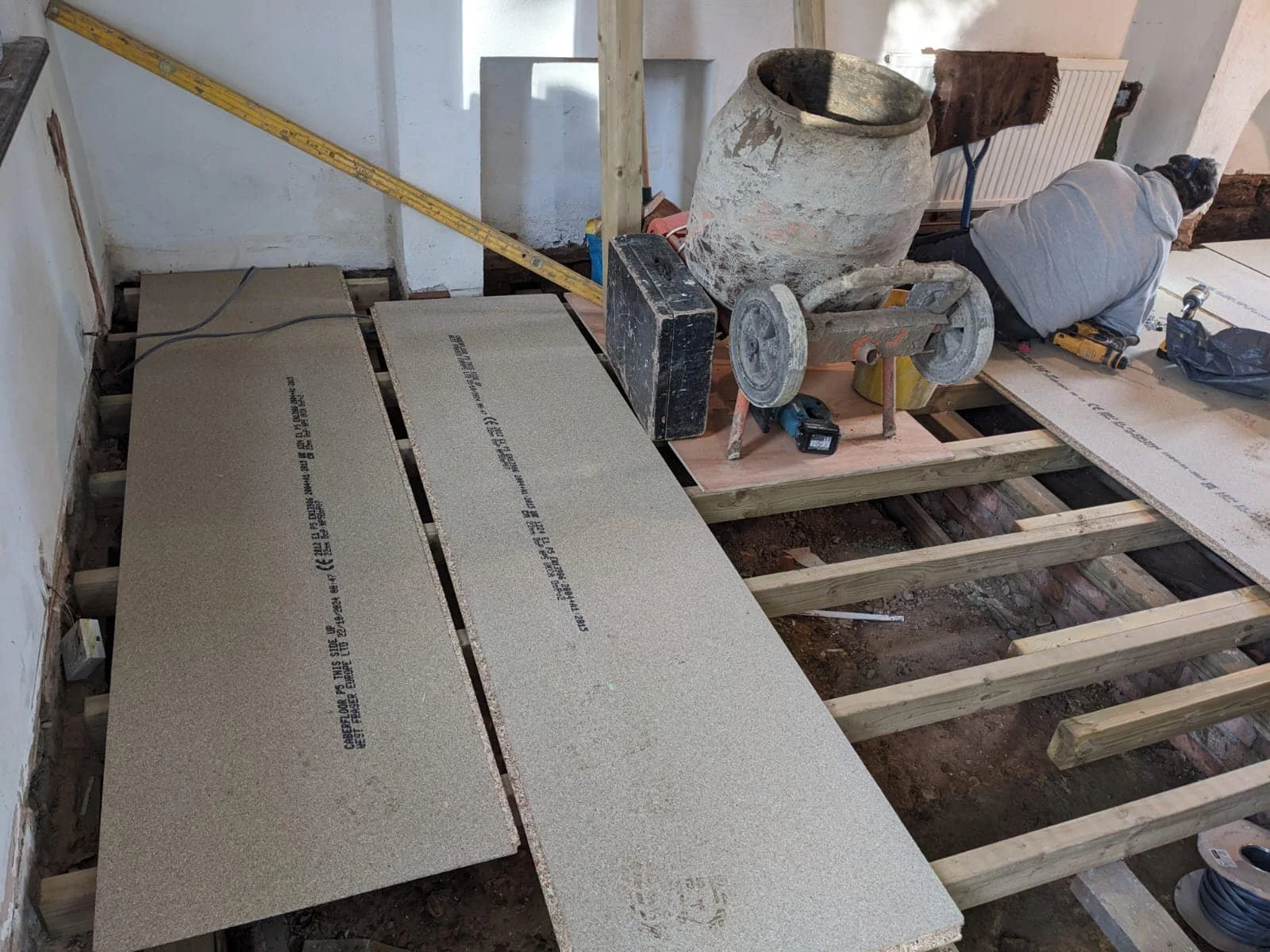 Construction worker installing new floor tiles in a room in north london for K G Croft. The image shows unfinished wooden floor joists, a concrete mixer, and tiles laid out on the floor.