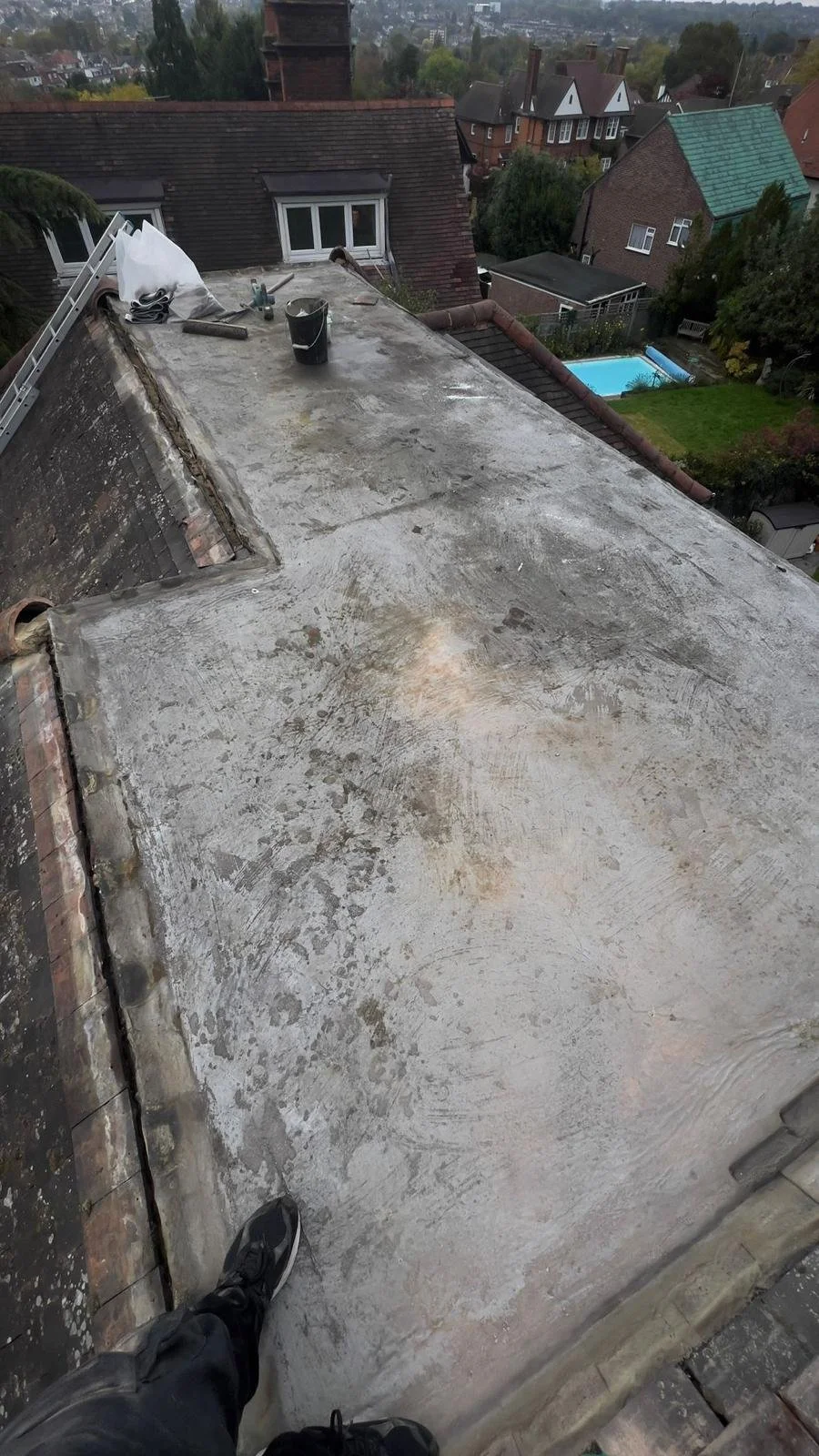 View from a rooftop in North London showing construction tools, a bucket, and a ladder, with neighbouring houses and a backyard with a swimming pool in the background. Renovation by K G Croft.