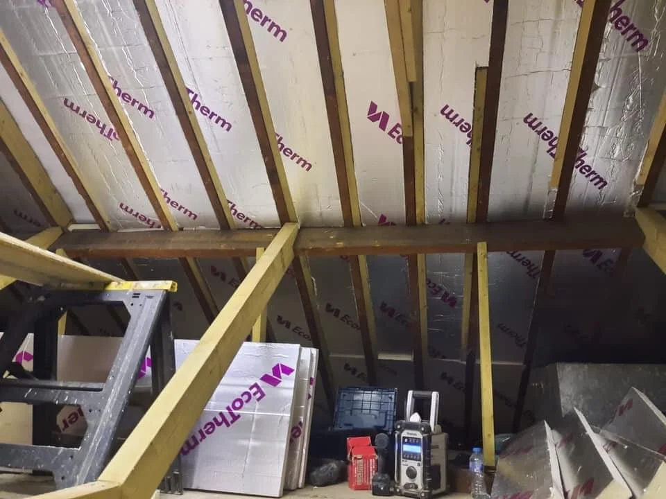 Interior view of a building under construction showing exposed wooden framing, insulation, and construction tools and materials on the floor.