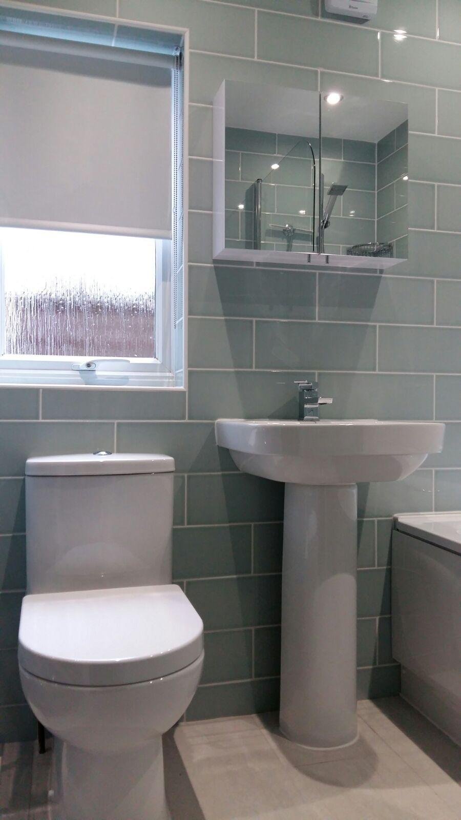A modern bathroom in north london with a white toilet, pedestal sink, mirror cabinet, window with a roller blind, and a washing machine, all against light green tiled walls. K G Croft offering bathroom solutions in north london.