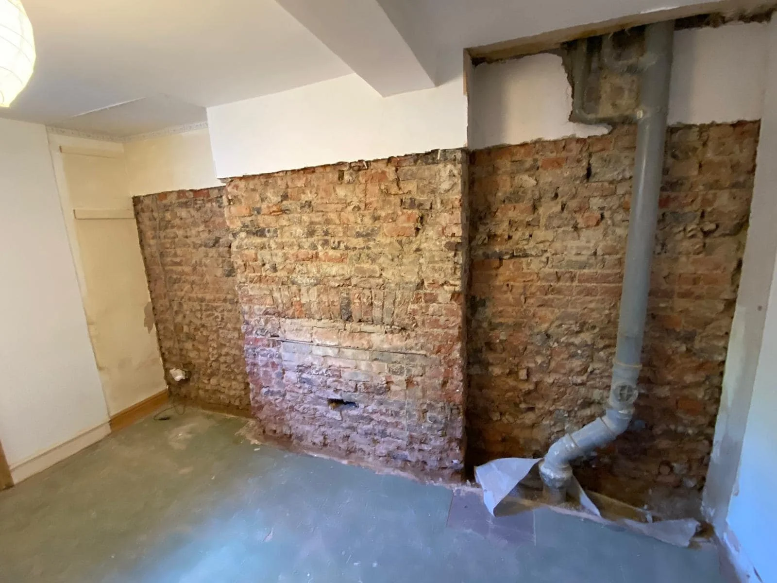 Partially demolished interior wall exposing brick and pipe plumbing in a room under renovation.