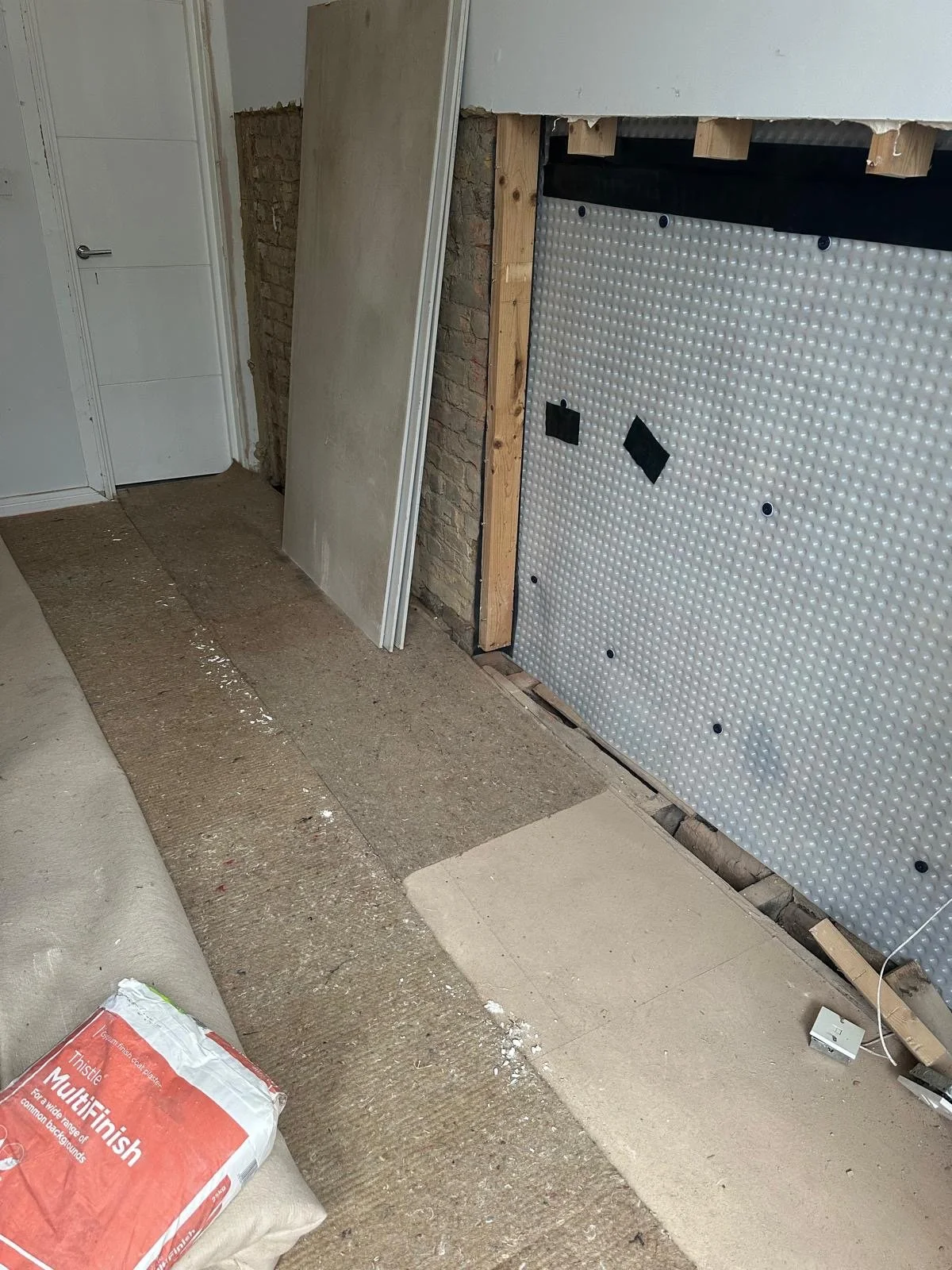 Room under renovation showing exposed drywall and brick wall, with construction materials and tools on the floor.